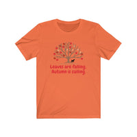 Image Description: Orange crew neck t-shirt. Orange and red maple leaves  on a brown tree trunk with a small black dog walking beside the tree trunk.  The www.DogsandTheirPaws.com url is under the tree.  The phrase, "Leaves are falling.  Autumn is calling." is below the image of the tree is in red.  