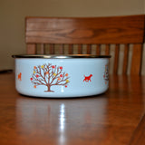 Autumn Is Calling Enamel Bowl - Image Description - White bowl with silver rim sitting on a wood table. The bowl shows autumn tree with red, orange and yellow paw prints as leaves with an orange pug on one side and  red dog on the other side of the tree.